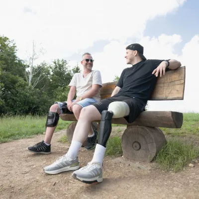 Zwei Männer mit Beinorthesen sitzen lachend auf einer Holzbank im Freien.