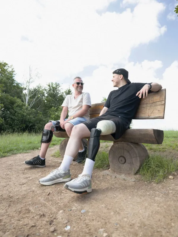 Zwei Männer mit Beinorthesen sitzen lachend auf einer Holzbank im Freien.