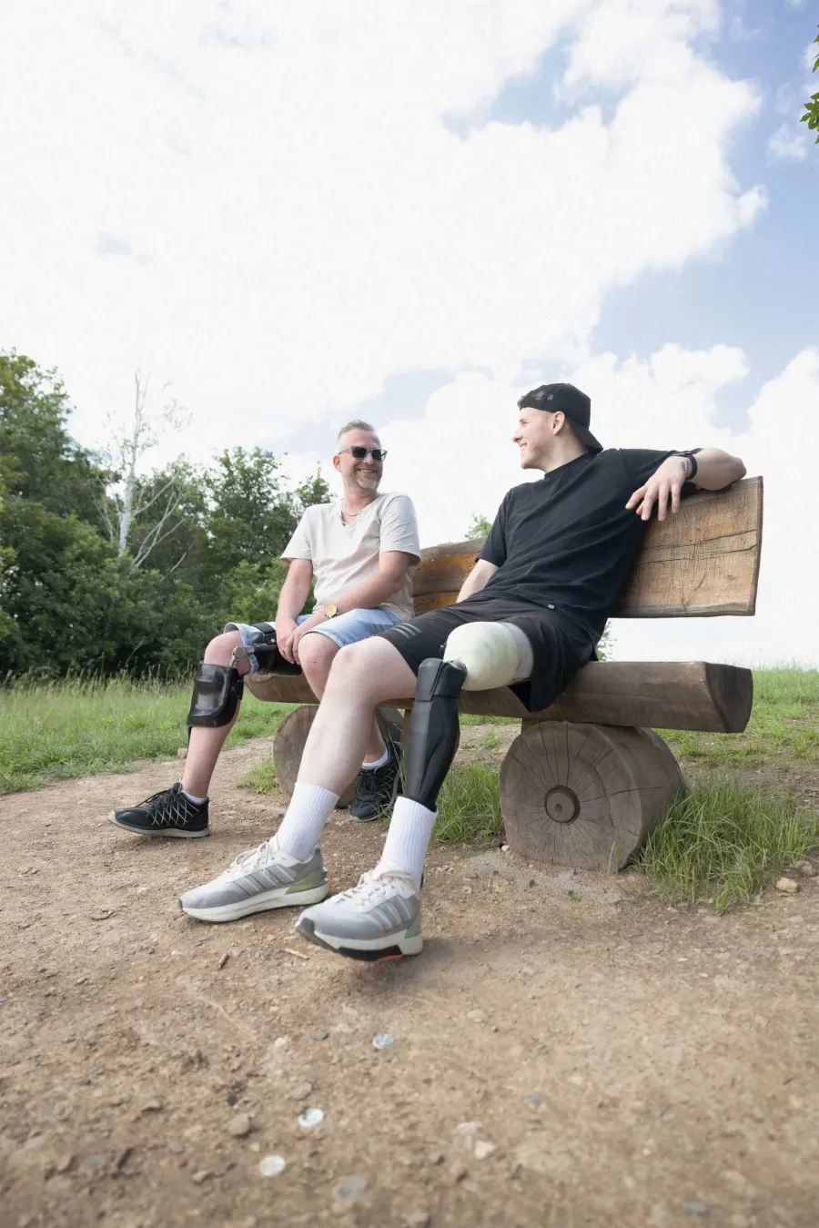 Zwei Männer mit Beinorthesen sitzen lachend auf einer Holzbank im Freien.