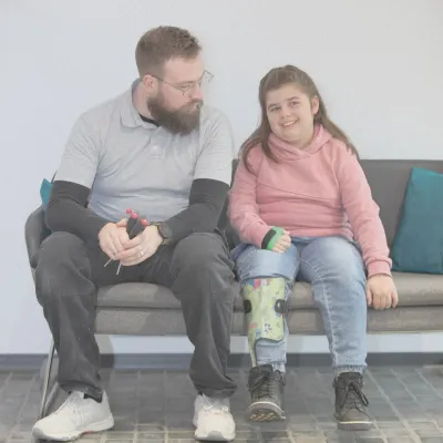 Mädchen mit bunter Beinorthese sitzt lächelnd neben Orthopädietechniker