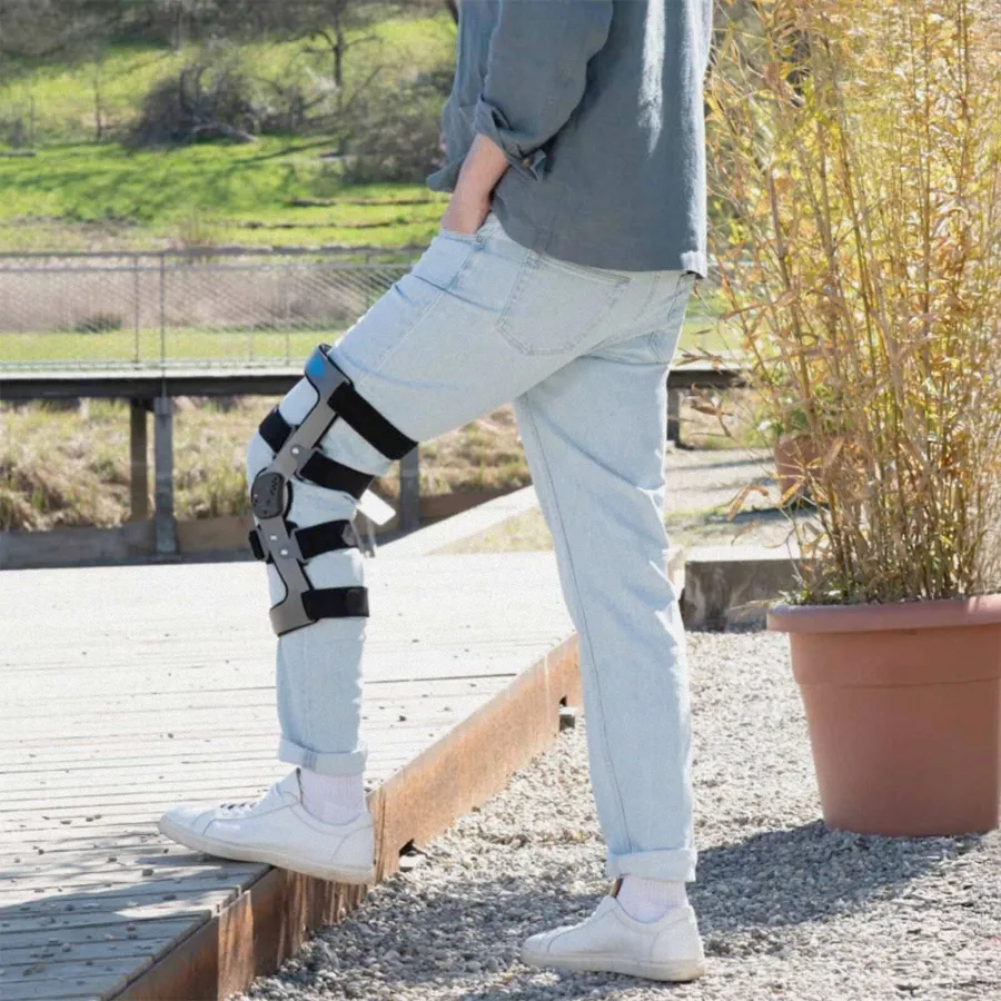 Person mit Knieorthese über der Jeans steht auf Holzsteg im Freien.